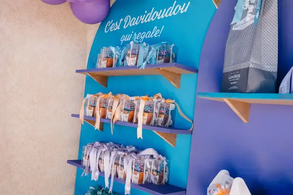 Planken met bedankjes in doorzichtige doosjes, in blauw en lila, onder een ballonboog